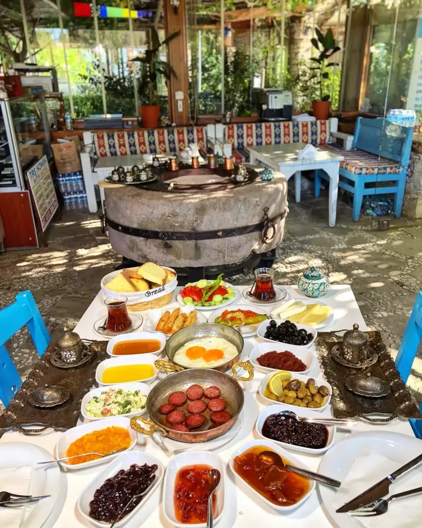 Geniş bir Taş Konak serpme kahvaltı masasında, sahanda sucuklu yumurta, zeytin çeşitleri, peynirler, rengarenk ev yapımı reçeller, salata ve taze ekmeklerle dolu tabaklar, geleneksel çay bardakları ve işlemeli kahve takımlarıyla birlikte, taş dokulu, yarı açık ve bitkilerle çevrili otantik bir mekan atmosferinde iştah açıcı bir görünüm sunuyor.