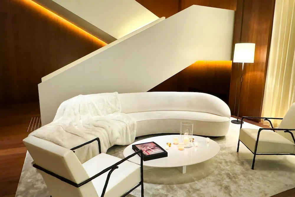 Modern lüks salon dekorasyonunda ahşap duvar panelleri, gizli LED aydınlatma, kavisli krem kanepe ve farklı dokularla oluşturulan katmanlı ve derinlikli iç mekân tasarımı.