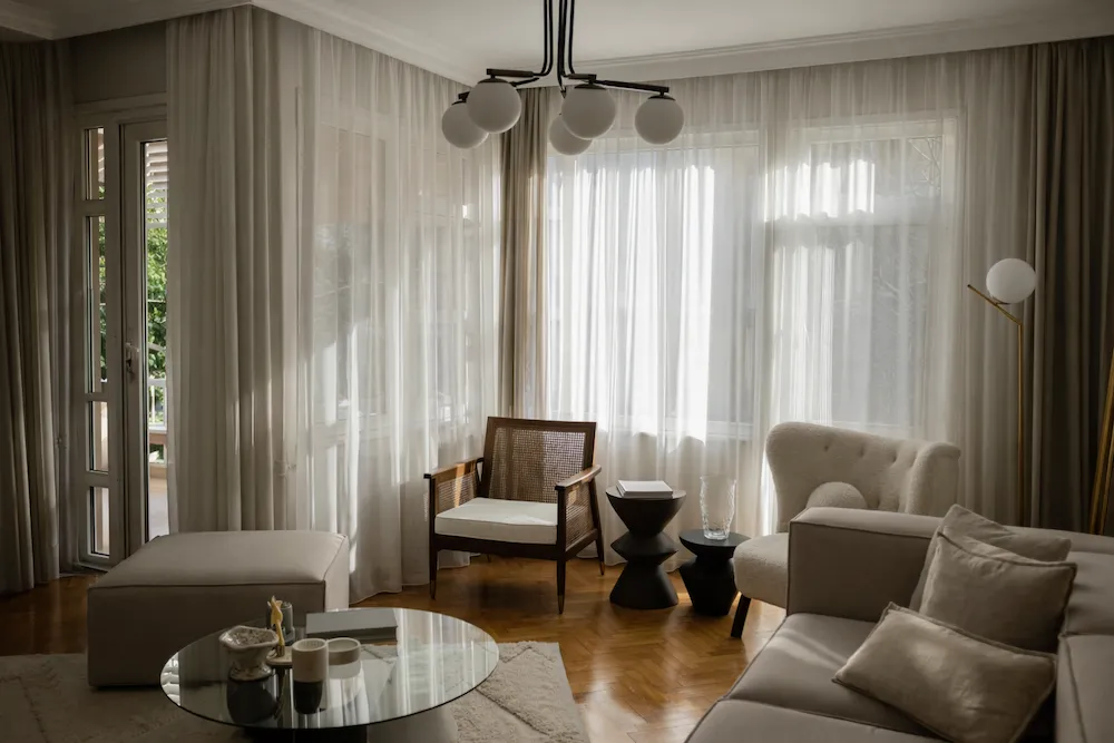 Lüks dekorasyonda doğal ışığa göre konumlandırılmış mobilya ve objeler; tül perdelerden süzülen gün ışığıyla aydınlanan modern salon, dengeli koltuk yerleşimi ve sıcak ahşap zemin detaylarıyla rafine iç mekân tasarımı.