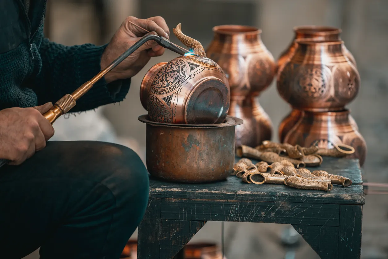 Mardin'in dar sokaklarında rastlanabilecek zanaatkarlık atölyelerinden birinde, el oymalı geleneksel desenlerle süslü bakır bir objeyi, alevli bir alet kullanarak işleyen, koyu renkli kazak giymiş bir ustanın elleri ve dizleri net bir şekilde görünüyor; arka planda da tamamlanmış diğer bakır eşyalar ve küçük işleme parçaları, şehrin popüler hediyeliklerinden olan bakır işlemelerin özgünlüğünü vurguluyor.