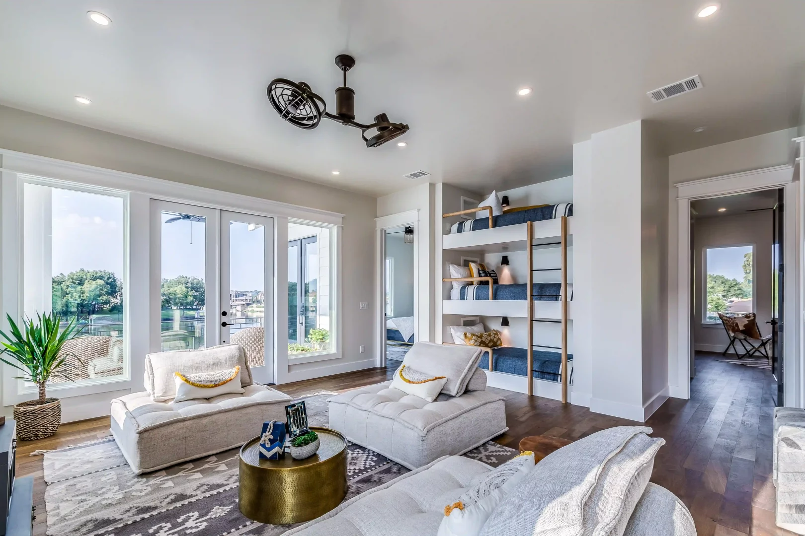 Bu modern Airbnb evinin geniş ve aydınlık oturma odasında, cam kapılardan görünen yeşil ağaçlar ve sakin bir göl manzarası eşliğinde, açık renkli, modüler kanepeler ve yuvarlak, altın rengi bir sehpa dikkat çekiyor; duvar boyunca uzanan mavi beyaz çizgili nevresimli çok katlı ranzalar, misafirler için hem şık hem de pratik bir uyku alanı sunuyor.