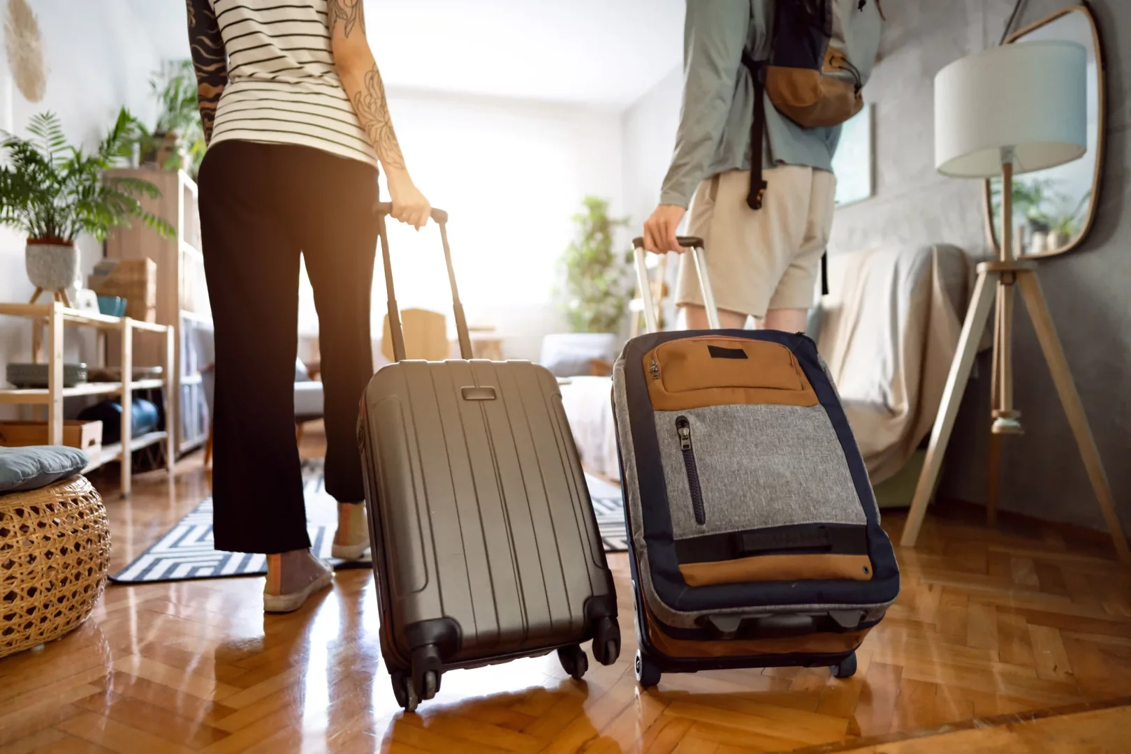 Güneşli bir ev ortamında, biri sırt çantalı, kolları dövmeli iki kişi, tekerlekli valizlerini çekerek Airbnb konaklamalarına giriş yapıyor veya çıkış yapıyor gibi görünüyor, bu durum seyahat ve geçici ev temalarını pekiştiriyor.