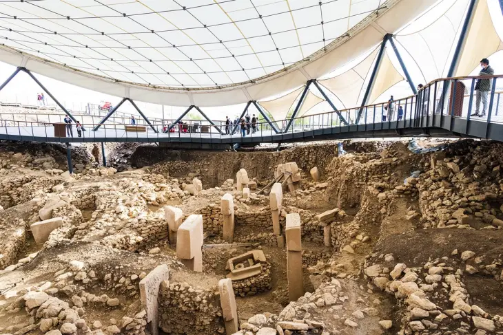 Göbeklitepe'deki modern koruyucu çatı altında sergilenen arkeolojik kazı alanında, T biçimli megalitik dikili taşlar ve antik yapılar toprağın içinden yükselirken, üst platformda ziyaretçiler bu görkemli tarihi kalıntıları dikkatle gözlemliyor.