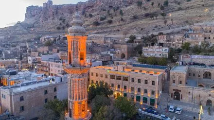 Akşamüstü çekilmiş bu havadan görünümde, ışıklandırılmış tarihi bir minare, hemen yanında modern bir butik otel olan Izala Butik Otel ve arka planda kayalık dağlarla çevrili eski bir şehrin büyüleyici manzarası gözüküyor.