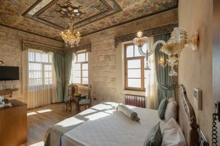 Çiçek desenli ahşap tavanı, taş duvarları ve kristal avizeleriyle geleneksel mimariyi modern detaylarla birleştiren, geniş pencerelerden süzülen güneş ışığıyla aydınlanan bu ferah butik otel odası, dinlendirici bir atmosfer ve zarif bir konaklama deneyimi sunuyor.
