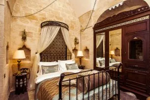 Mardius Otel'in taş duvarlı ve kemerli tavanlı otantik bir odasında, ferforje detaylı cibinlikli yatak, ahşap oymalı aynalı gardırop ve yatak başı lambalarıyla sıcak bir atmosfer sunuluyor.