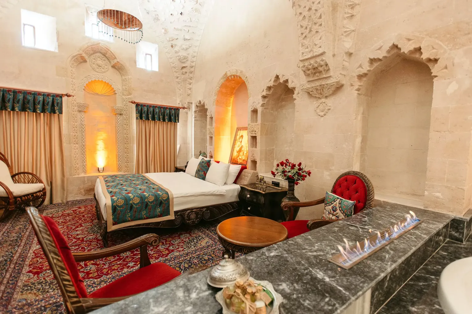 Bir Mardin otelinin geleneksel taş işçiliği ve kemerli nişlerle süslenmiş, desenli bir yatak örtüsüyle dikkat çeken büyük yatağa sahip, lüks bir konuk odası. Kırmızı halı, kadife detaylı koltuklar ve modern bir şömine bu otantik atmosferi tamamlıyor.