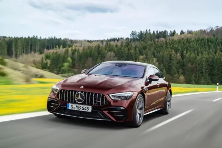 Koyu bordo renkli, sportif hatlara sahip Mercedes-AMG GT 4 Kapı Coupe, yeşil ağaçlar ve sarı çiçeklerle kaplı tepelerin çevrelediği virajlı yolda hızla ilerleyerek performans odaklı yapısını gözler önüne seriyor.