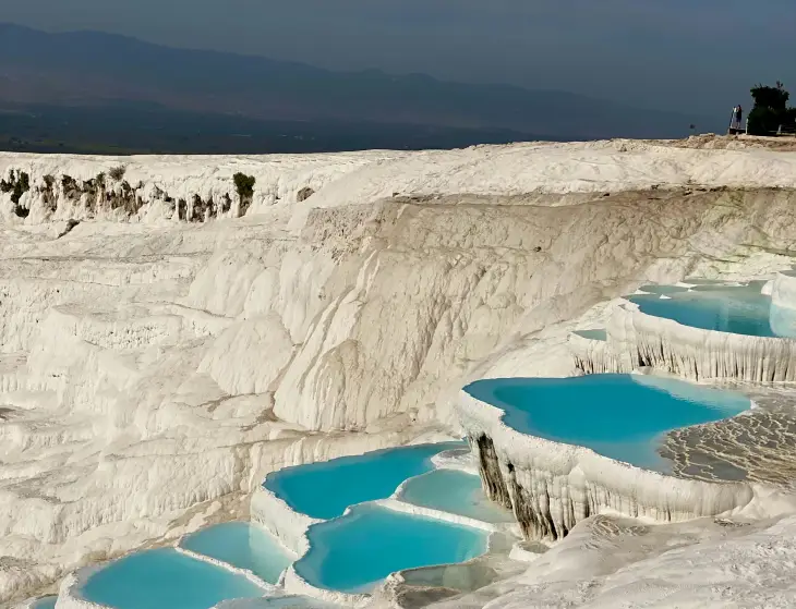Türkiye'nin Denizli ilindeki Pamukkale'de, kireçtaşı travertenler üzerinde oluşan göz alıcı turkuaz renkli termal havuzların ve bembeyaz basamaklı oluşumların geniş bir manzarasını gösteren büyüleyici bir fotoğraf.