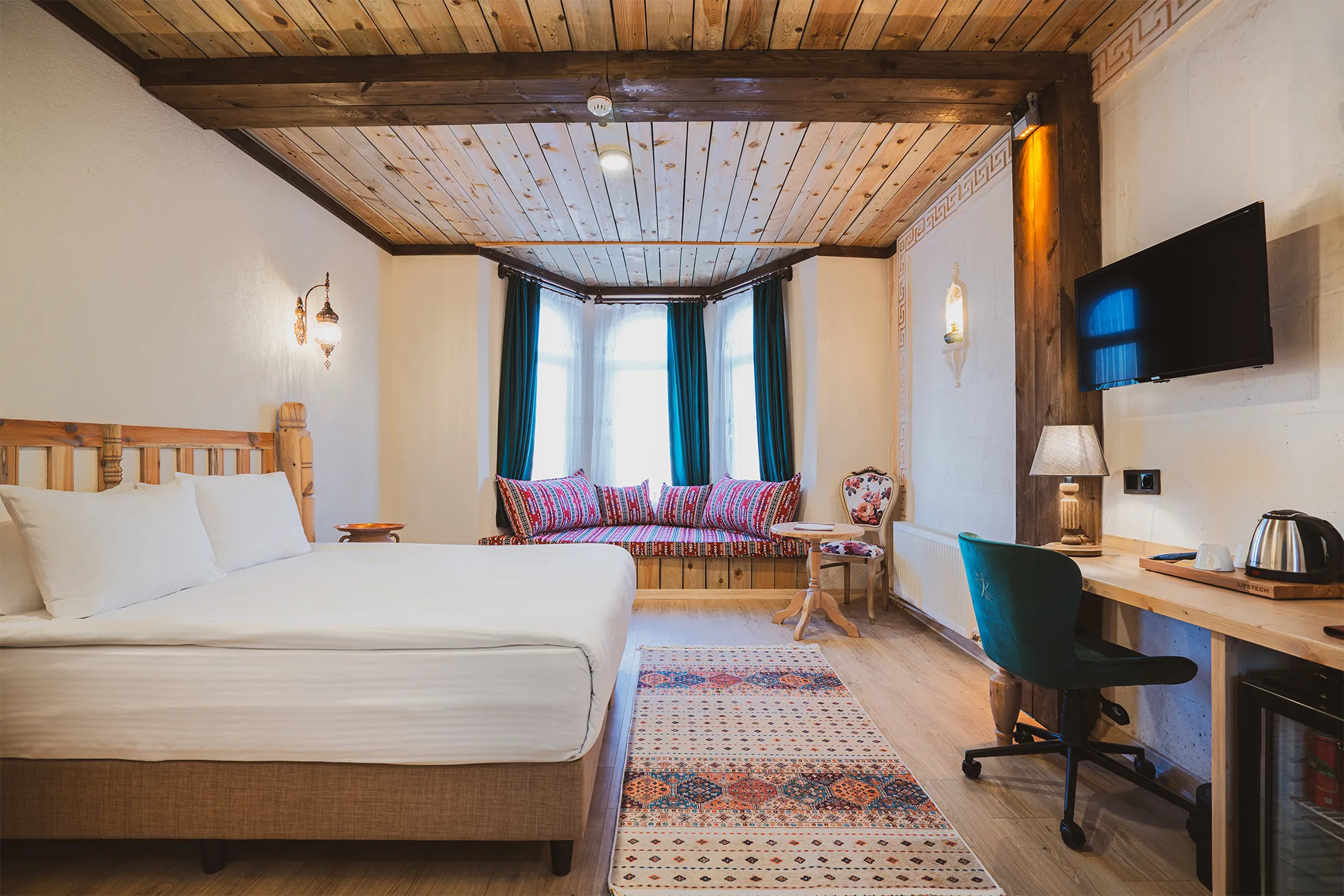 Ahşap tavan ve kirişlerle tasarlanmış, geleneksel ile modern unsurları harmanlayan Raymar Hotels odası; beyaz nevresimli yatağı, renkli desenli minderlere sahip şık cumba penceresi, duvara monte TV ve çalışma masasıyla misafirlerine konforlu bir ortam sunuyor.
