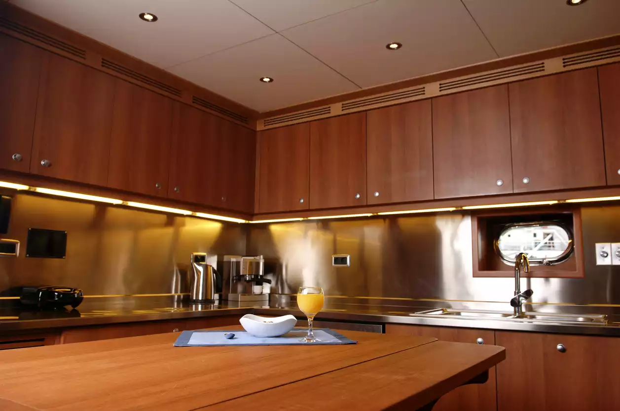 Ahşap dolapları, paslanmaz çelik çalışma yüzeyleri ve tezgah altı LED aydınlatmalarıyla modern bir tasarıma sahip lüks yat mutfağında, lavabonun üzerinde oval bir gemi penceresi ve ön plandaki ahşap masada bir bardak portakal suyu ile beyaz bir kase dikkat çekiyor.