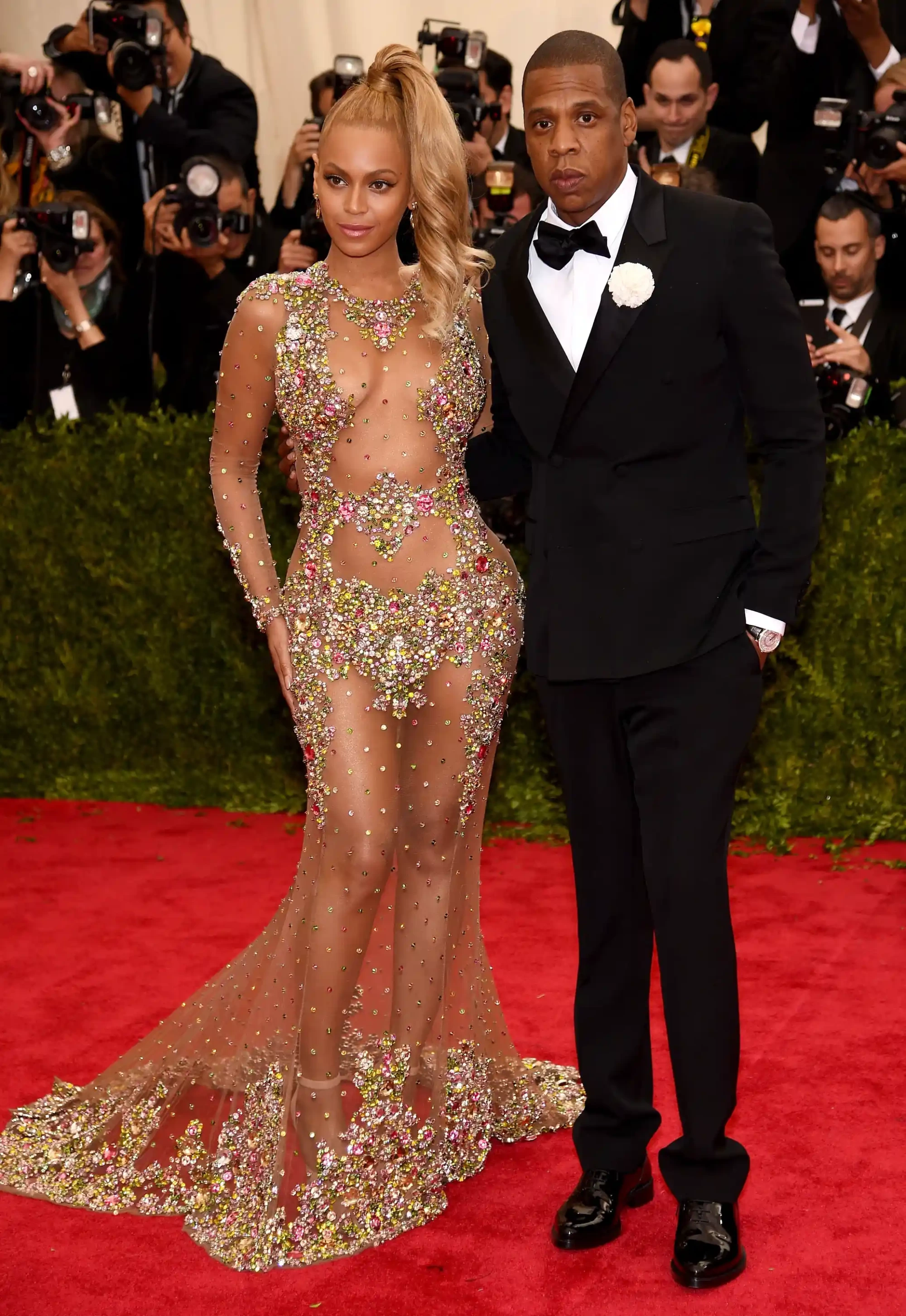 2015 Met Gala’da kırmızı halıda poz veren Beyonce, sıkı at kuyruğu ve çok renkli kristallerle bezeli, transparan, uzun kollu, yere kadar uzanan iddialı Givenchy elbisesiyle dikkat çekerken, Jay-Z de klasik siyah smokiniyle eşlik ediyor.
