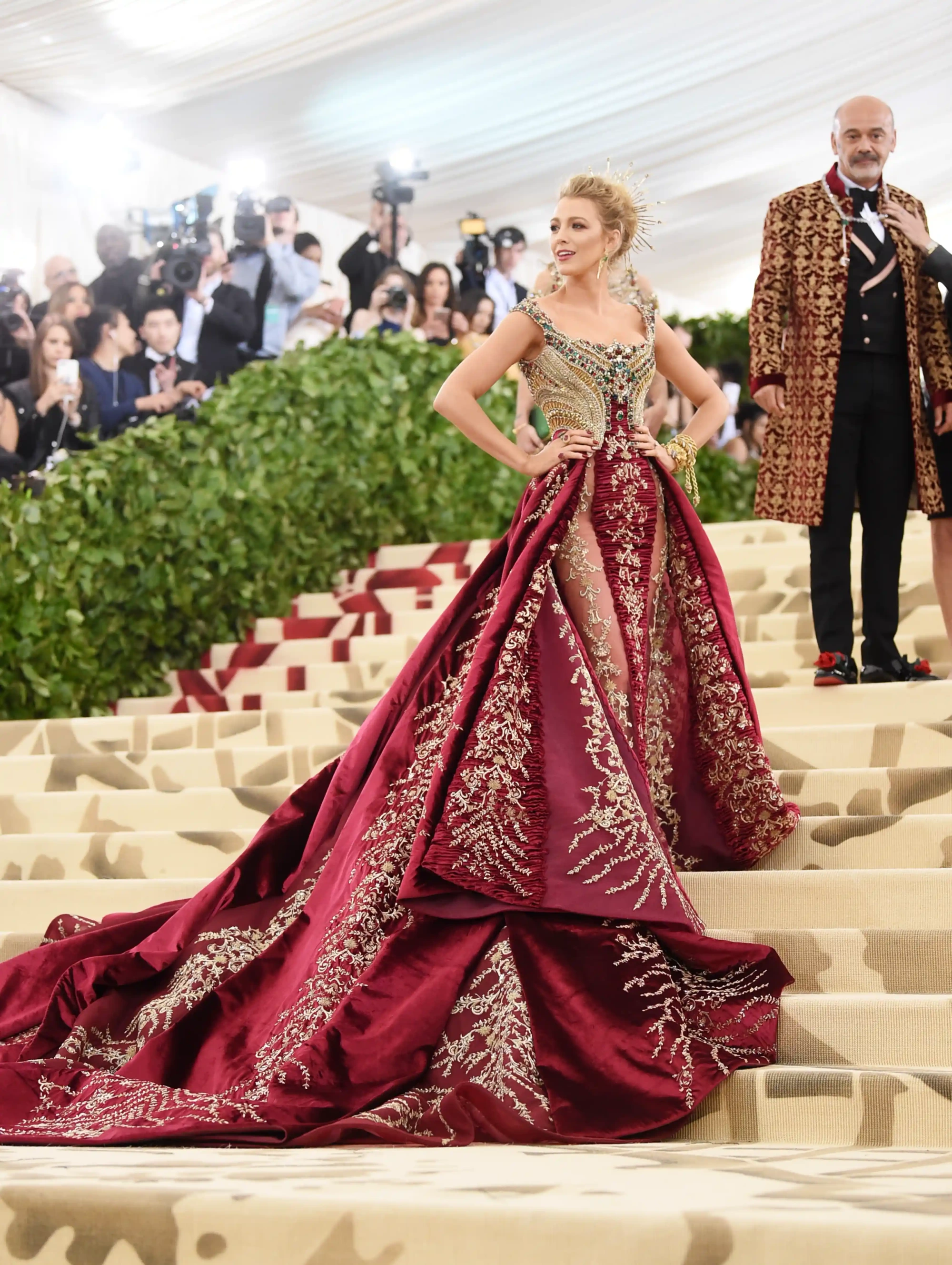 Blake Lively, 2018 Met Gala etkinliğinde, bordo ve altın işlemelerle süslü, uzun kuyruklu gösterişli Versace elbisesiyle büyük bir merdivende dramatik bir poz verirken görülüyor, zarif tacı ve mücevherleri ise bu ikonik Met Gala görünümünü tamamlıyor.