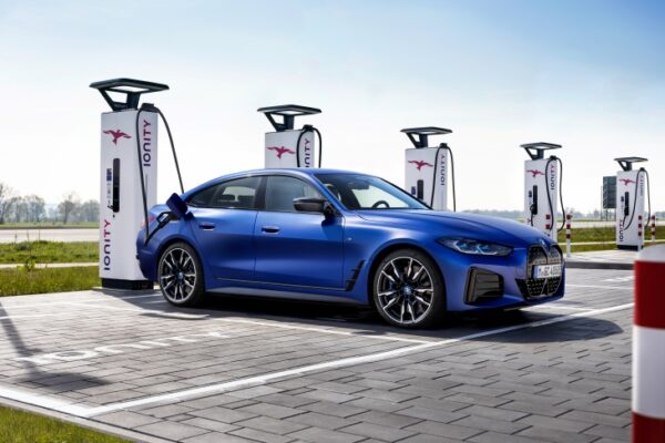 Parlak mavi renkli bir Yeni BMW i4, Ionity logolu beyaz şarj ünitelerinin önünde, arka tarafına bağlı şarj kablosuyla tamamen elektrikli bir otomobil olarak hızlı şarj imkanlarını ve elektrikli sürüş deneyimine geçişi vurgulayarak bekliyor.