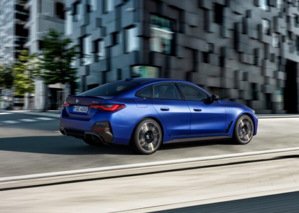 Mavi renkli Yeni BMW i4, modern mimariye sahip şehir binalarının önünden yüksek hızla ilerlerken, tamamen elektrikli bu şık otomobilin sportif ve dinamik hatlarını arka çeyrek açıdan etkileyici bir şekilde sergiliyor.