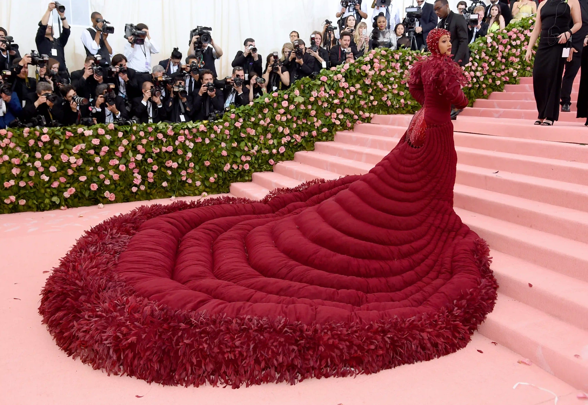 Cardi B, pembe merdivenlerde, kabarık, koyu kırmızı, kapitone görünümlü ve etekleri tüy detaylı devasa bir kuyruğa sahip elbisesi ve uyumlu başlığıyla 2019 Met Gala'da objektiflere poz verirken, arkasındaki güllerle süslü duvarın arkasından çok sayıda fotoğrafçı onu görüntülüyor.