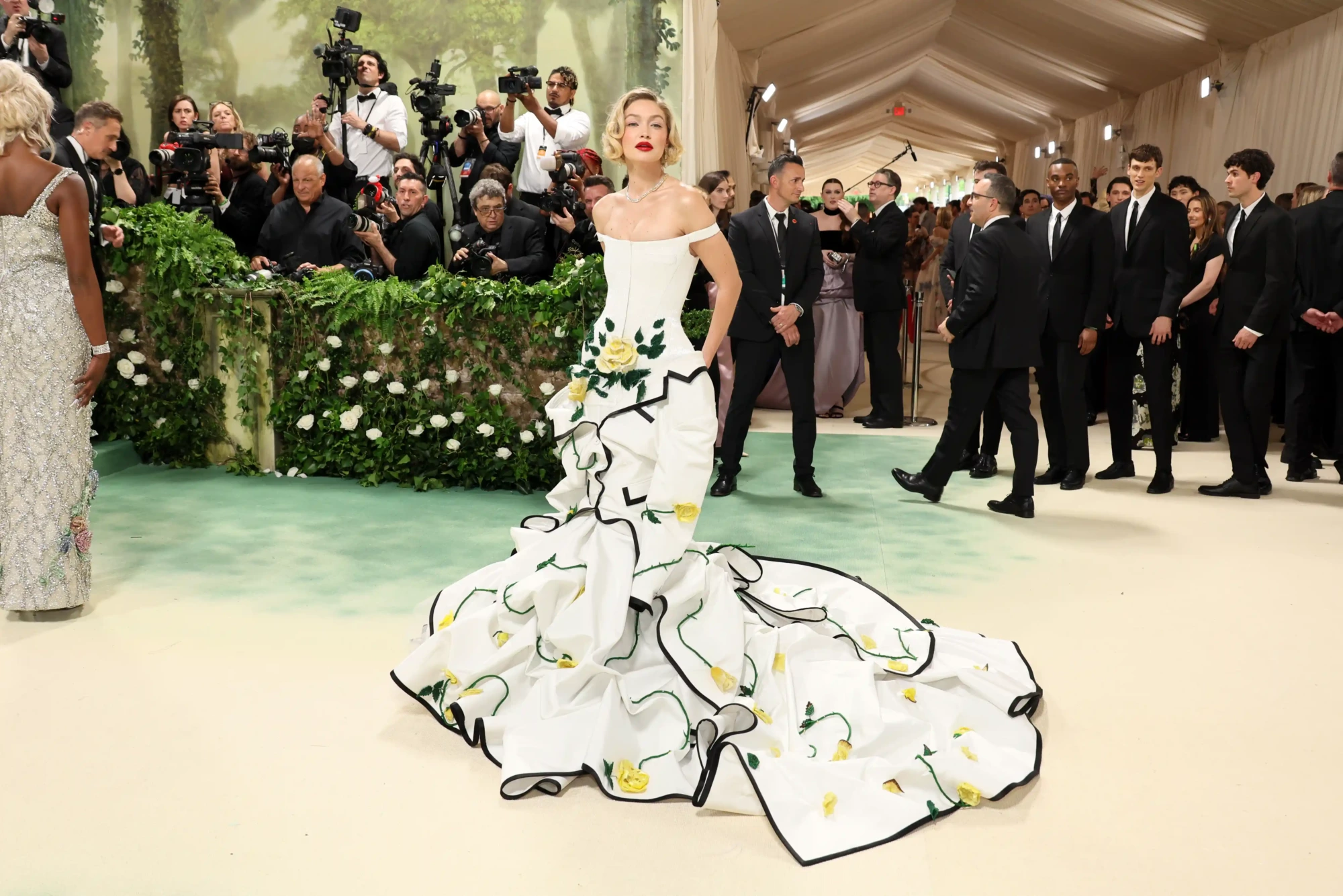 Gigi Hadid, moda dünyasının Oscar’ı olarak bilinen Met Gala’da, sarı gül işlemeli ve siyah biyeli, straplez, kabarık etekli beyaz bir elbiseyle kameralara poz vererek ikonik görünümler sergiliyor.