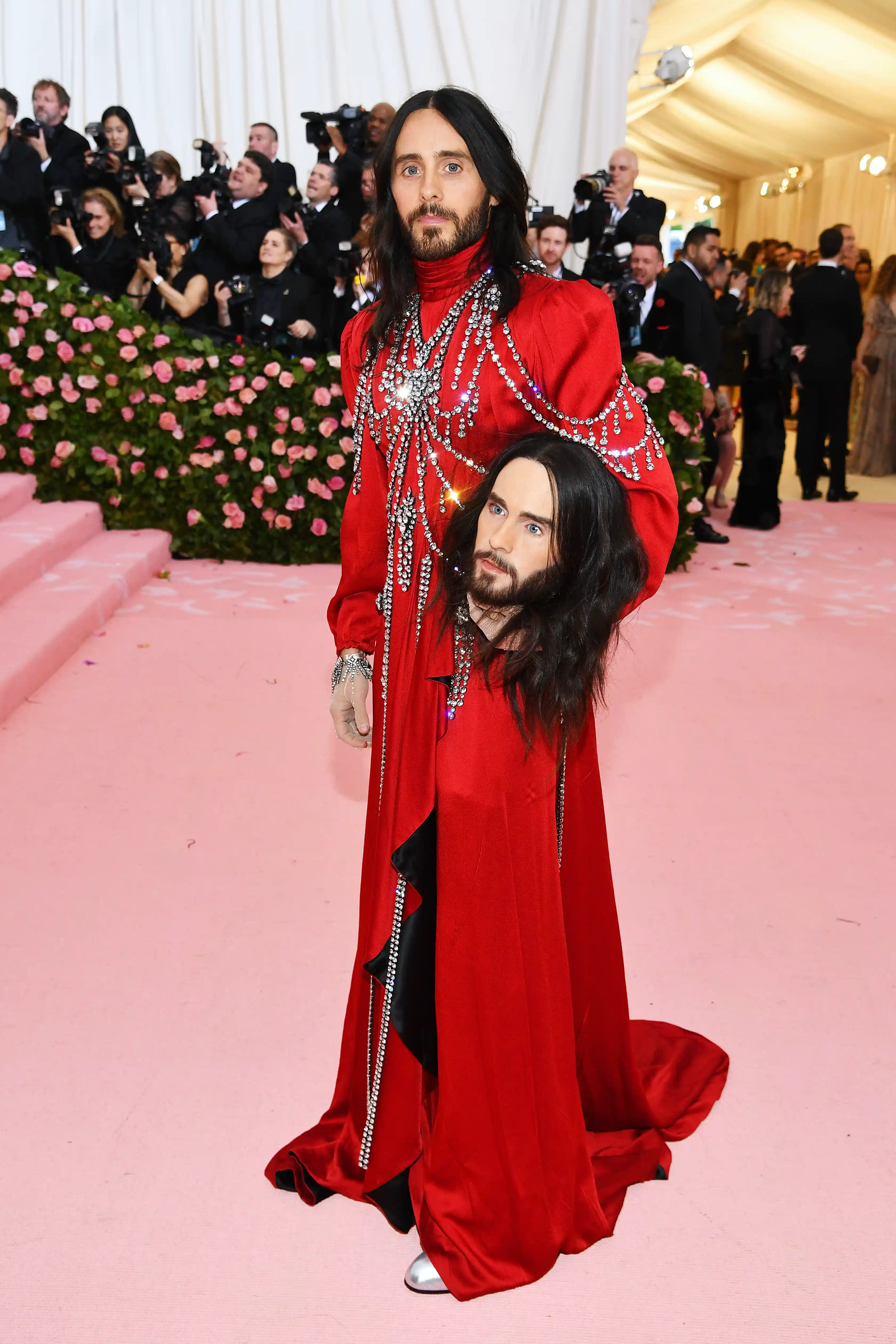 Jared Leto, 2019 Met Gala kırmızı halısında, uzun koyu saçları ve sakalıyla, parlak gümüş taşlarla süslü kırmızı elbisesi içinde, kendi kafasının bir replikasını kolunda taşıyarak tüm dikkatleri üzerine çekiyor ve gecenin ikonik görünümleri arasında yerini alıyor.