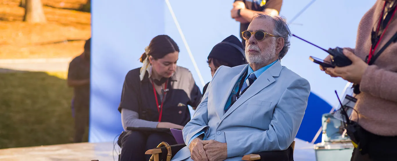 Güneş gözlüklü, beyaz sakallı yaşlı bir adam, açık mavi takımıyla açık havada bir film setinde veya prodüksiyon alanında, tabletine bakan bir set görevlisi ve telsiz tutan başka bir ekip üyesiyle çevrili olarak sandalyede oturuyor.