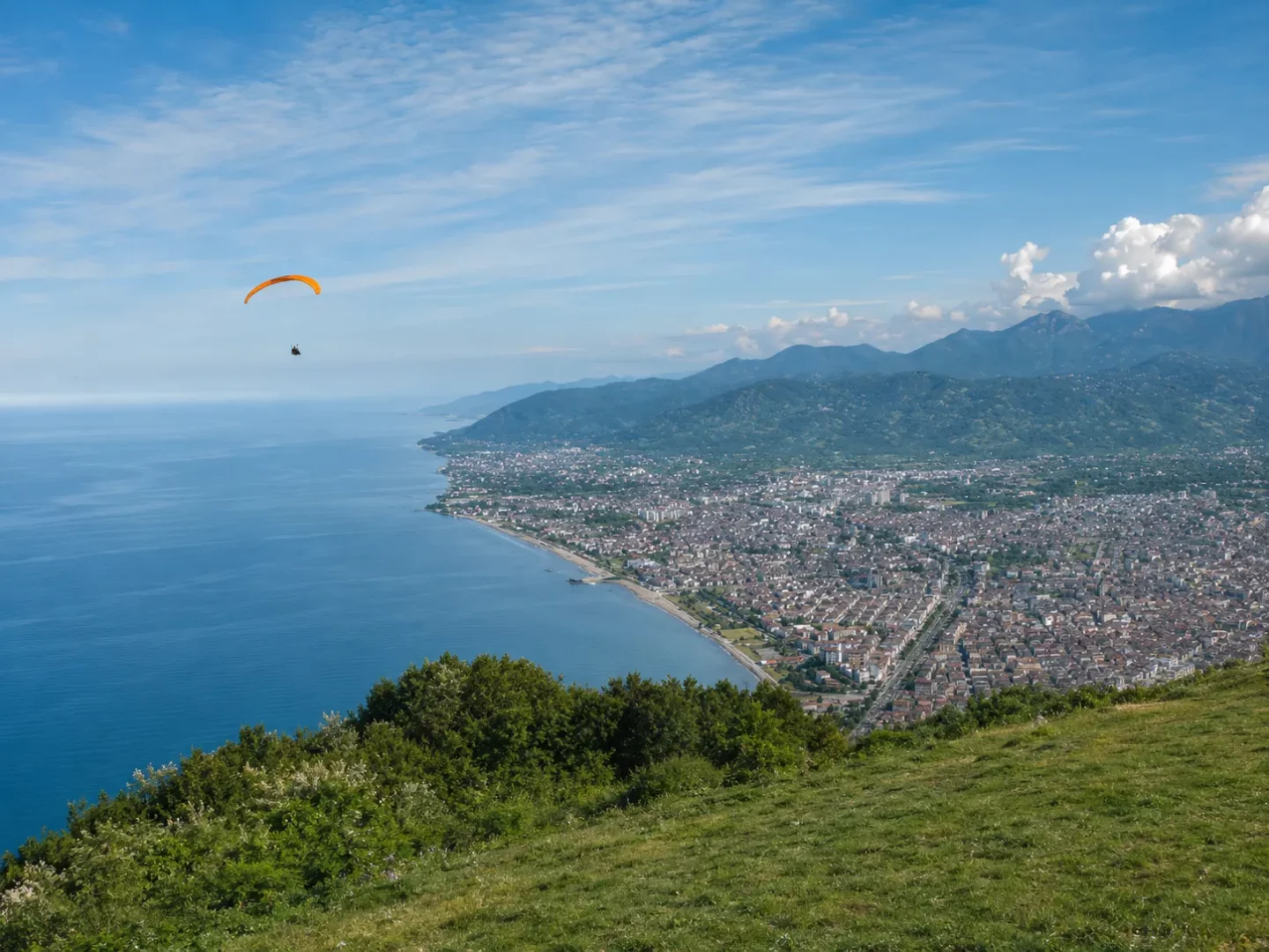 Ordu Boztepe'den Karadeniz'in masmavi suları ve kıyı şeridi boyunca uzanan Ordu şehir merkezinin geniş bir kuşbakışı görünümünde, güneşli bir günde açık mavi gökyüzünde parlak turuncu bir paragliding süzülüyor.