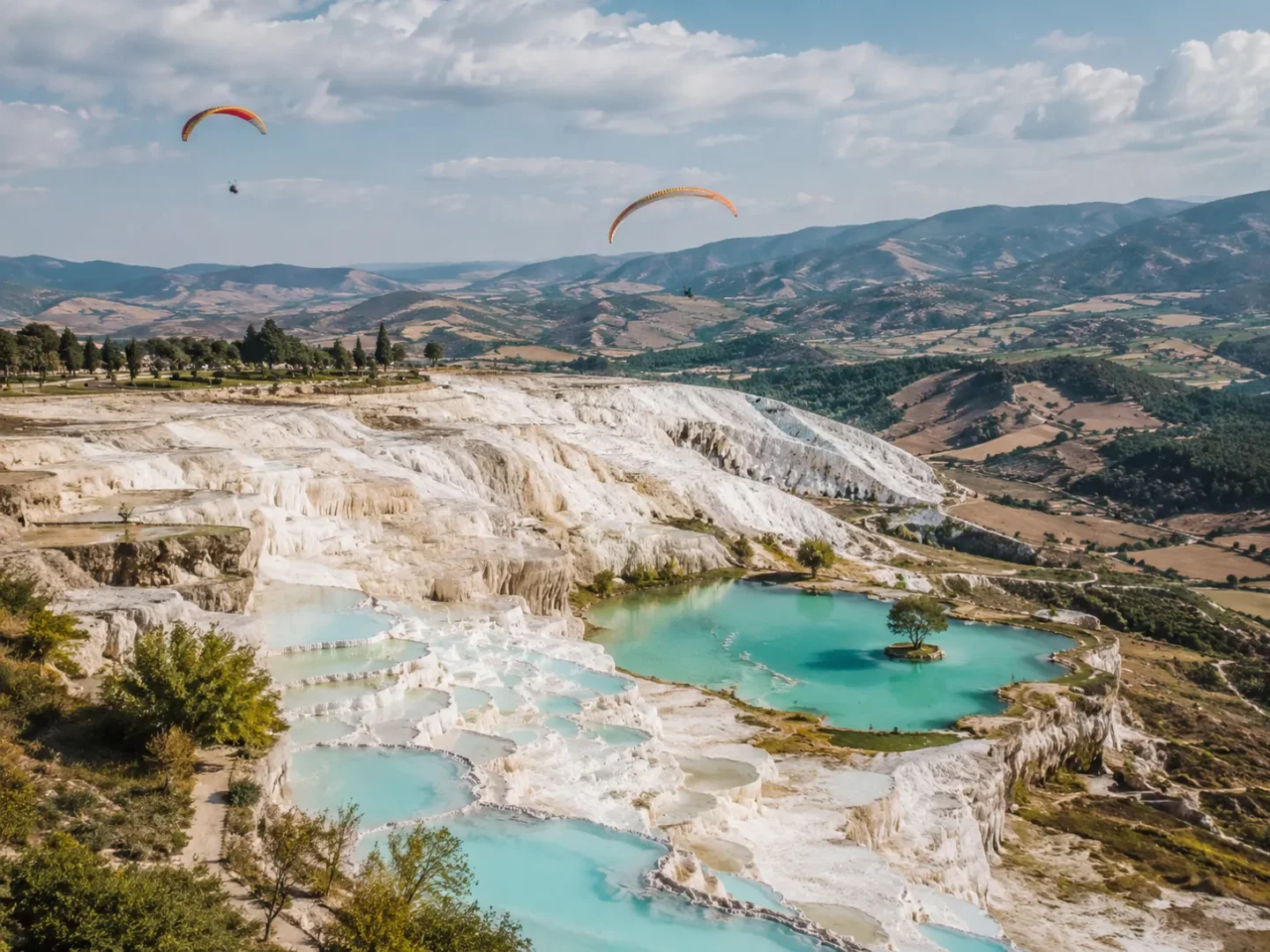 Pamukkale'nin eşsiz beyaz travertenleri ve turkuaz sularının üzerinde süzülen renkli yamaç paraşütleri, yemyeşil tepelerle çevrili bu doğal cennetin Dinamit Tepesi'nden görünen büyüleyici manzarasını gözler önüne seriyor.