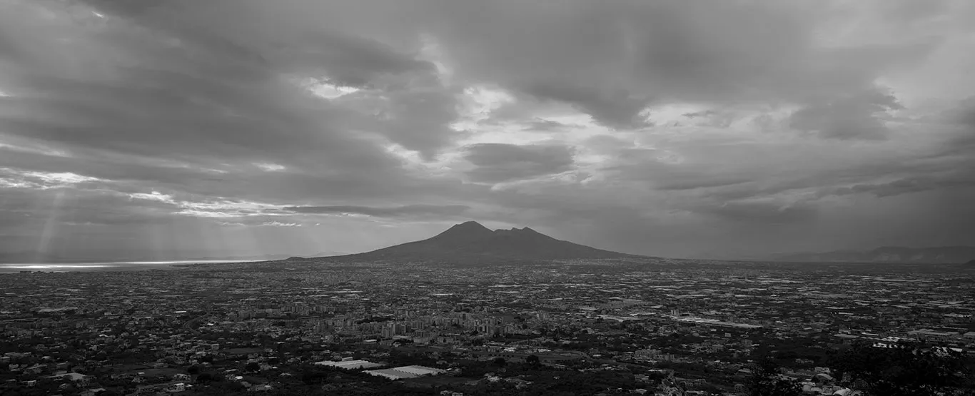 Bu siyah beyaz panoramik fotoğrafta, yoğun bulutların altındaki Pompei ve çevresini kapsayan geniş bir şehir manzarası, uzakta heybetli Vezüv Yanardağı ve sol yanda güneş ışınlarının aydınlattığı denizle birlikte dramatik bir gökyüzünün altında seriliyor.