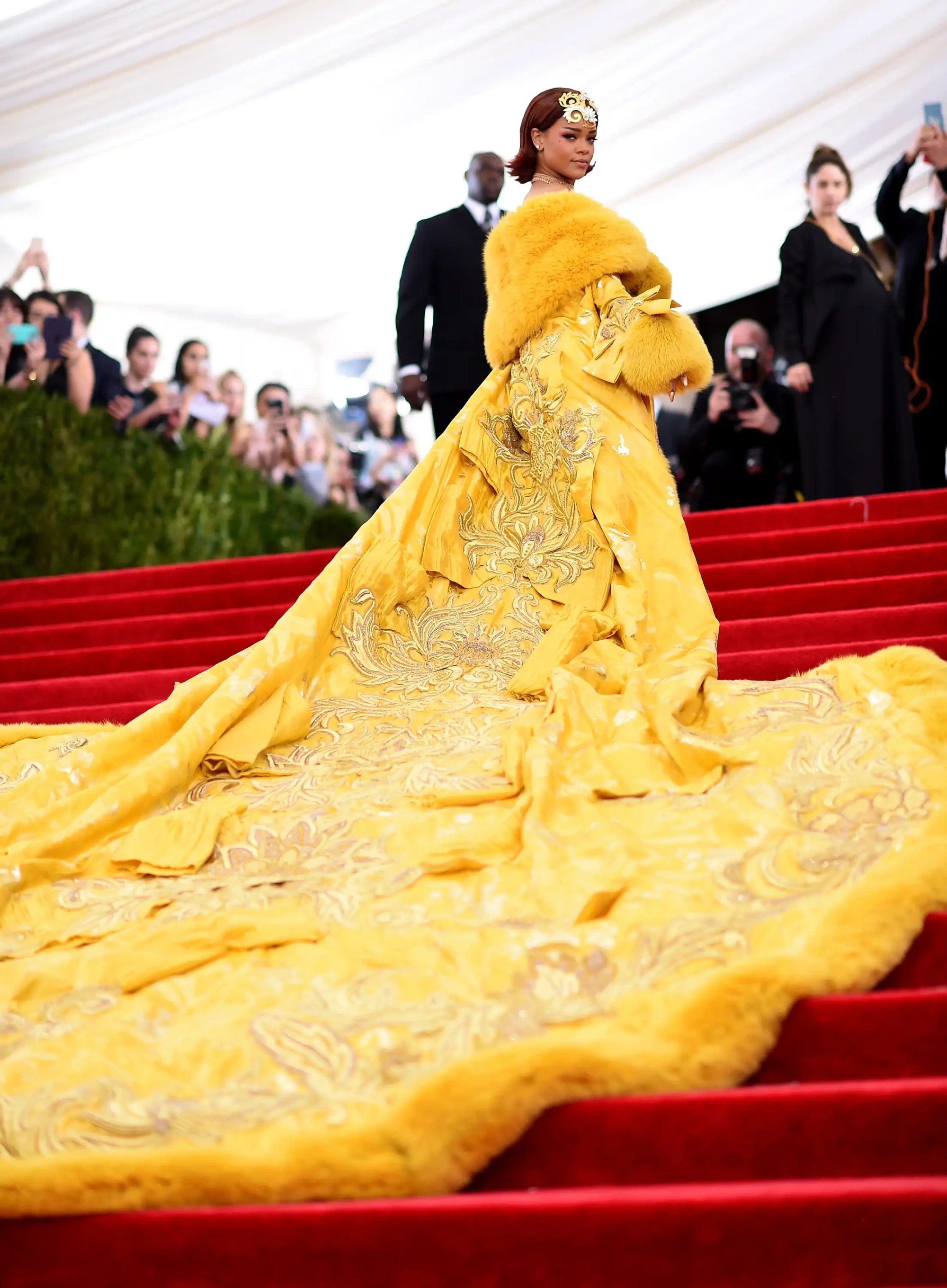 Met Gala'nın kırmızı halısında, omuzlarında sarı bir kürk şal ve başında altın bir taçla poz veren Rihanna, etekleri altın işlemelerle bezeli, uzun ve kabarık sarı elbisesiyle modanın en büyük gecesinin en ikonik görünümlerinden birini gözler önüne seriyor.