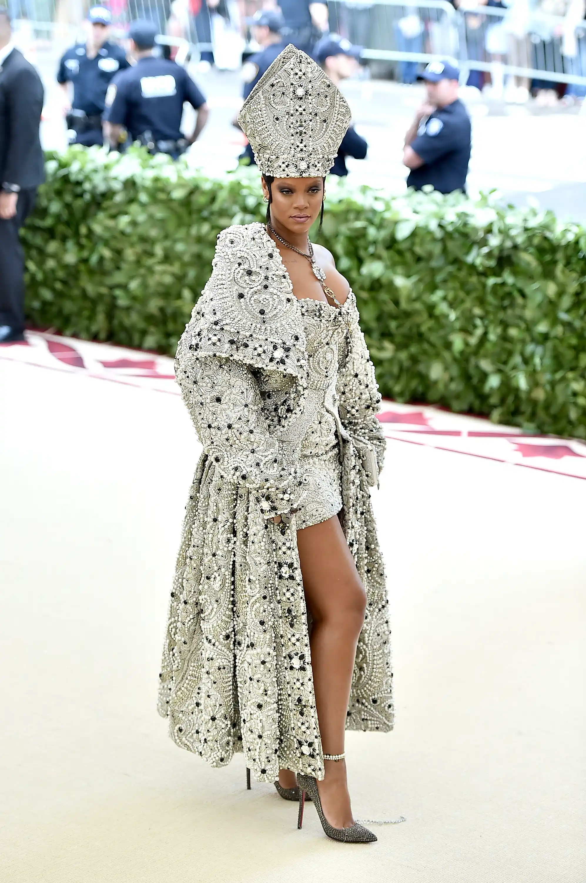 Rihanna, 2018 Met Gala için Katoliklikten ilham alan, baştan aşağı inci ve siyah taşlarla işlenmiş mitre şapkası ve pelerinli mini elbisesiyle kırmızı halıda dururken, bu ikonik görünümü Christian Louboutin ayakkabıları ve zarif takılarla tamamlanıyor.