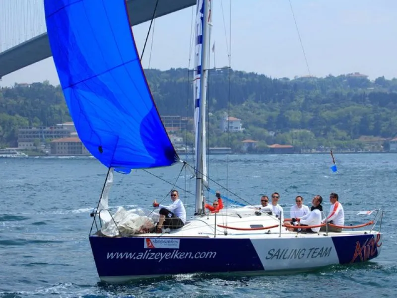 Alize Yelken Okulu'nun mavi ana yelkeni açık bir yelkenlisi, beyaz tişörtlü bir grup yelkenciyle İstanbul Boğazı'nda ilerlerken, güneşli havada köprülü ve yeşil tepeli manzarayla bütünleşiyor.