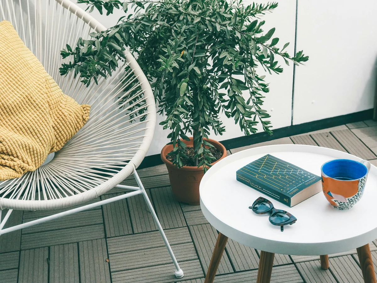 Beyaz Acapulco tarzı bir balkon koltuğu üzerinde sarı minder, yanında yeşil yapraklı saksı bitkisi ve yuvarlak beyaz bir sehpanın üzerinde koyu renkli bir kitap, güneş gözlüğü ve renkli bir kupa ile huzurlu ve modern bir balkon dekorasyonu.