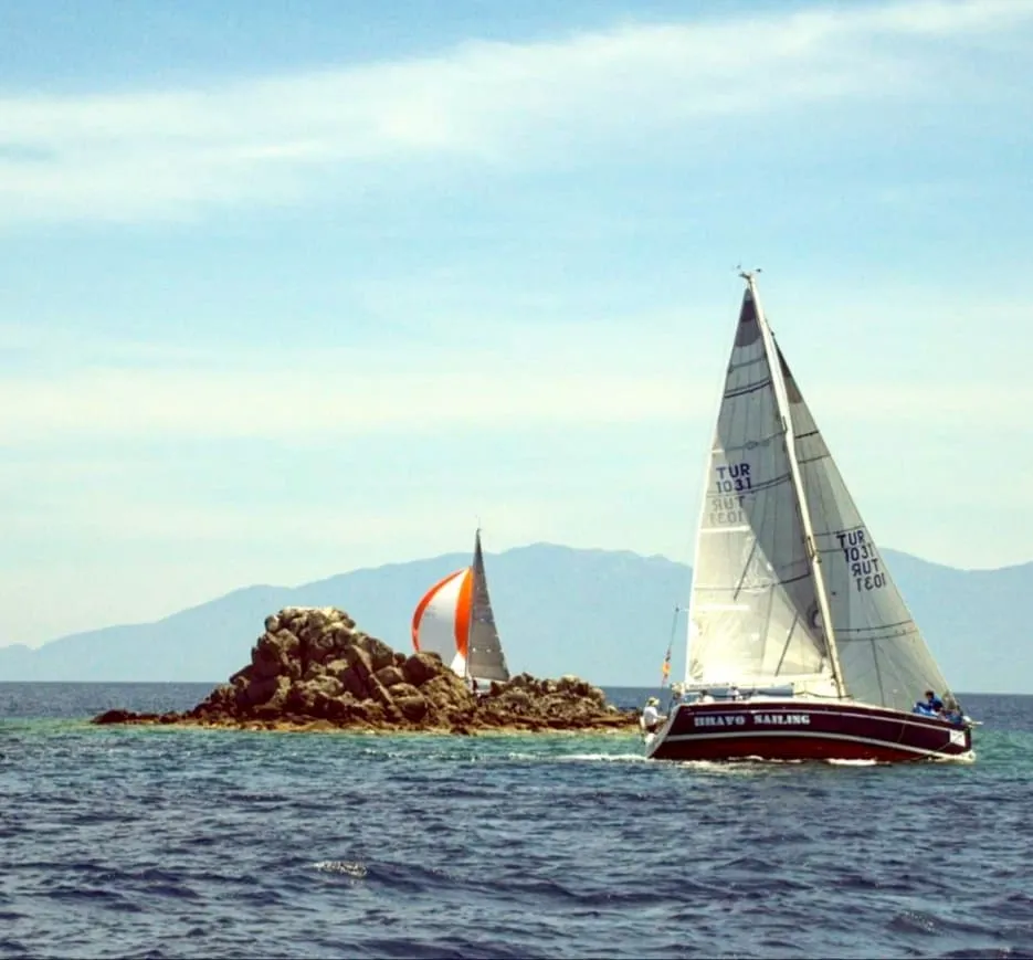 Berrak mavi gökyüzü ve uzak dağ manzarası eşliğinde dalgalı denizde süzülen, bordo gövdeli ve beyaz yelkenli BRAVO SAILING teknesi ile kayalık adacığın ardında turuncu-beyaz yelken açmış bir diğer tekne, Bodrum'da yelken okulu deneyiminin sunduğu eşsiz güzelliği ve öğrenme coşkusunu vurguluyor.