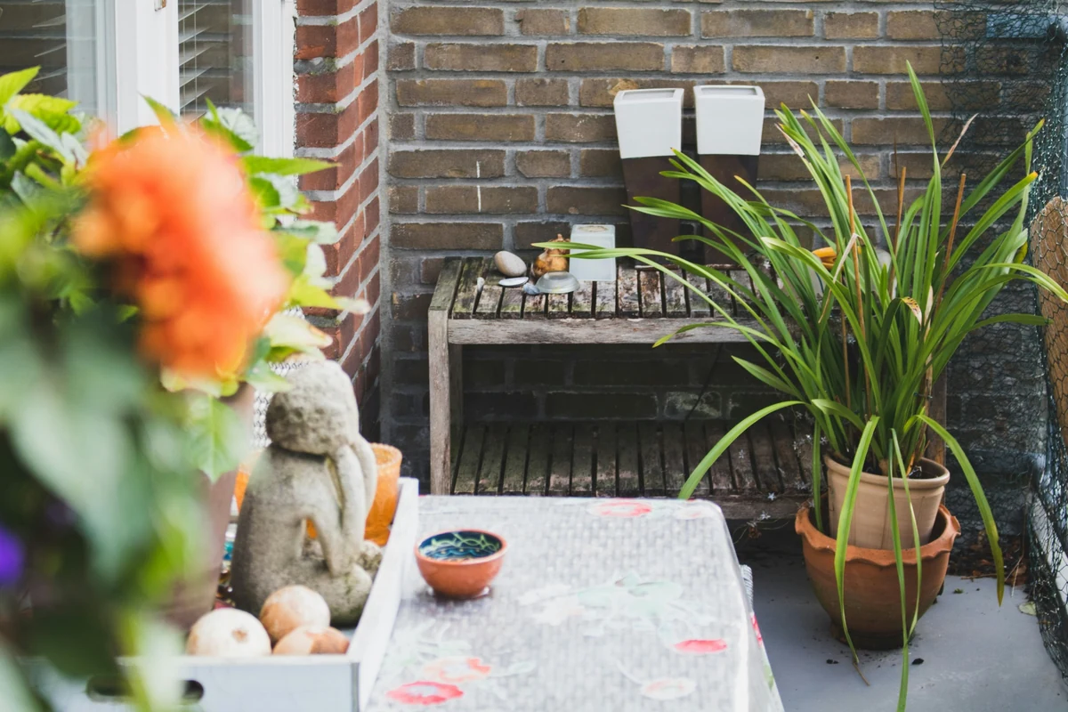 Tuğla duvarın önündeki ahşap raf üzerinde taşlar ve mumlukların bulunduğu bu modern balkonda, duvara monte edilmiş iki beyaz saksı ve zeminde iç içe geçmiş büyük toprak saksılar içinde canlı yeşil yapraklı bir bitki ile şık bir atmosfer yaratılmış.