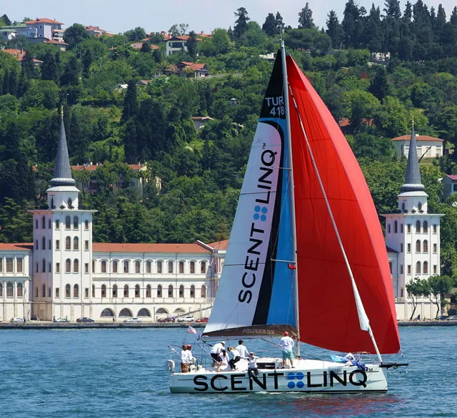 Scentlinq logolu kırmızı ve beyaz yelkenleriyle üzerinde öğrencilerin veya ekibin bulunduğu yelkenli tekne, İstanbul Boğazı'nda kuleli tarihi binalar ve yemyeşil tepeler önünde Cenoa yelken okulu derslerini sergiliyor.
