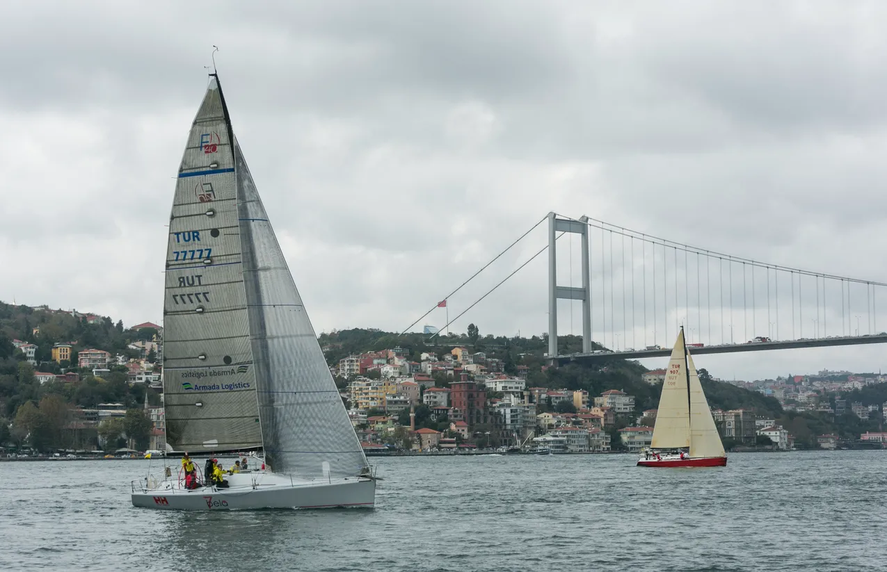 İstanbul Boğazı'nda gri bir havada, üzerinde TUR 77777 numarası ve Armada Logistics yazılı büyük bir yelkenli teknenin, Bosphorus Köprüsü ve yemyeşil yamaçlardaki binalar önünde, daha küçük kırmızı gövdeli başka bir yelkenliyle birlikte ilerlediği bu sahne, İstanbul'da yelken sporunun canlılığını ve Boğaz'ın eşsiz güzelliğini yansıtmaktadır.