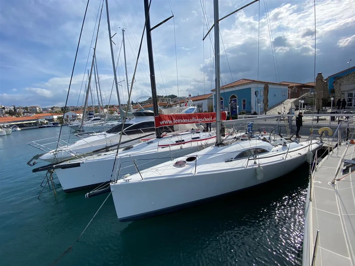 İzmir'deki bir marinada, Lemon Sailing School'a ait bembeyaz yelkenli tekneler sakin masmavi su üzerinde sıra halinde bağlı dururken, arka planda geleneksel mimarili binalar ve bulutlu gökyüzü, bu yelken okulu için huzurlu ve davetkar bir ortam oluşturuyor.