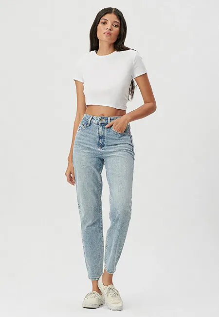 Açık mavi yüksek bel mom fit jean pantolon ve beyaz crop tişört giyen kadın model; beyaz spor ayakkabıyla stüdyo çekimi, düz açık gri arka plan.