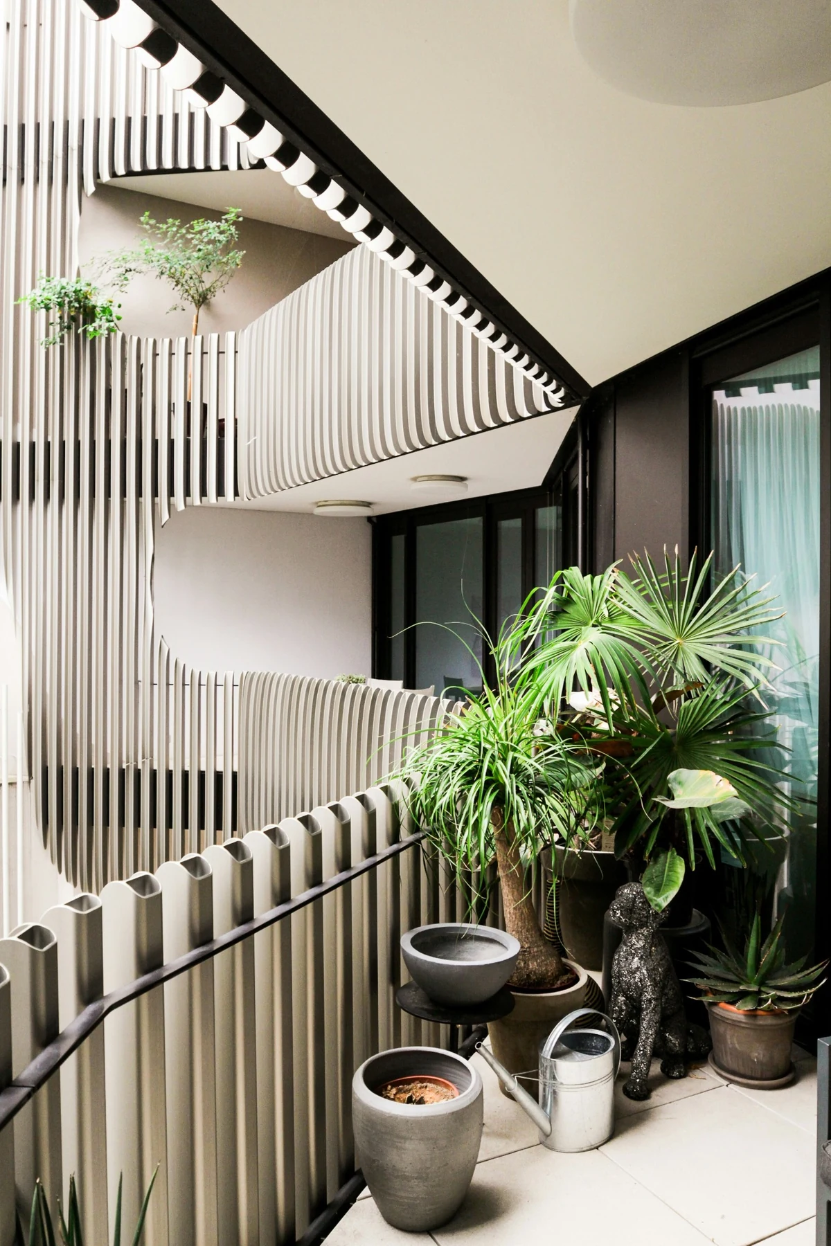 Bu fotoğrafta, dikey ve kıvrımlı panellerle oluşturulmuş modern balkon mimarisi, zemin ve üst katmanlarda yer alan çeşitli bitkiler, şık saksılar, bir sulama kabı ve dekoratif bir köpek heykeliyle estetik ve doğal bir yaşam alanı sunuyor.