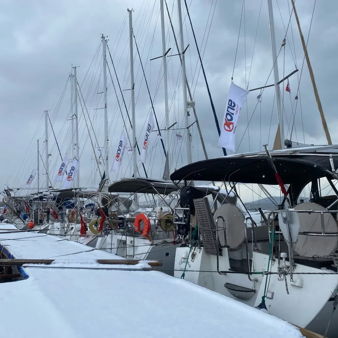 Karlarla kaplı bir iskelede sıralanmış, direklerinde beyaz one yacht logolu bayrakların dalgalandığı birçok yelkenli yat, bulutlu ve soğuk bir havada kış yatçılığının sakin ve etkileyici atmosferini yansıtıyor.