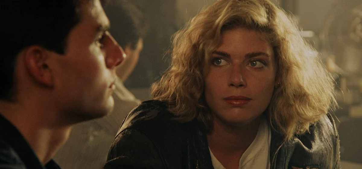 Top Gun Maverick filminden, sarı kıvırcık saçlı, açık renk gözlü, koyu renk bir ceket giyen Charlie'nin (Kelly McGillis) yüzünün belirgin olduğu ve kısmen görünen Maverick'in (Tom Cruise) profiliyle birbirlerine dikkatle baktıkları loş ışıklı bir sahne.