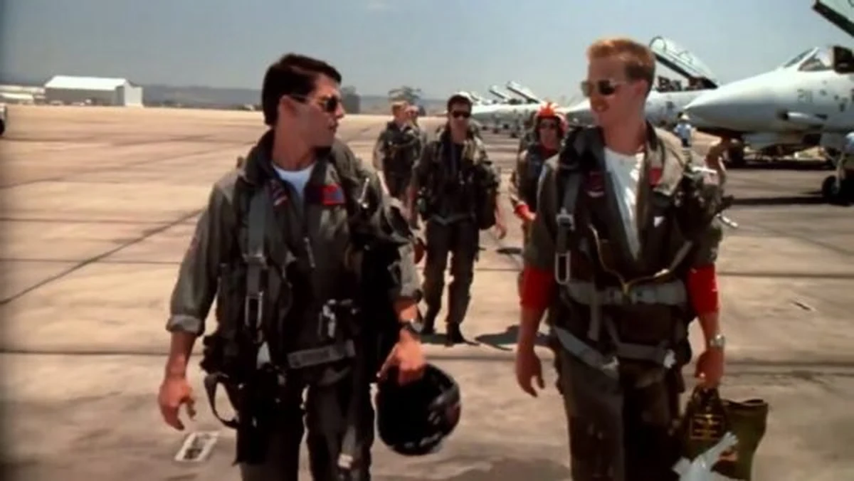 Maverick ve Iceman karakterleri, pilot tulumları ve havacı gözlükleriyle uçak pistinde yürürken, Maverick sol elinde miğfer, Iceman ise sağ elinde çanta taşırken görülüyor; arka planda ise park etmiş savaş uçakları ve diğer ekip üyeleri dikkat çekiyor.