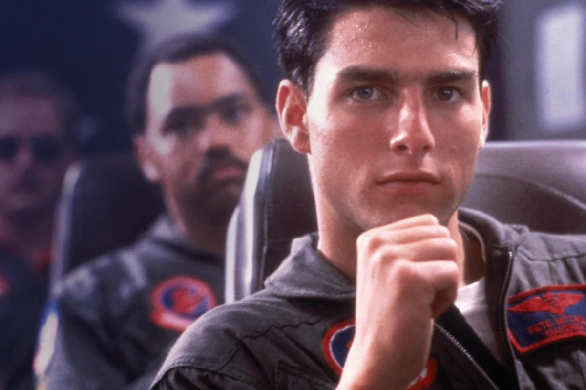 Top Gun Maverick filminden bir kokpit sahnesinde, genç Tom Cruise pilot tulumu içinde, sağ yumruğu çenesinin altında ciddi bir ifadeyle dururken, tulumundaki Maverick isimli yama dikkat çekiyor ve arkasında bulanık görünen iki kişi daha bulunuyor.