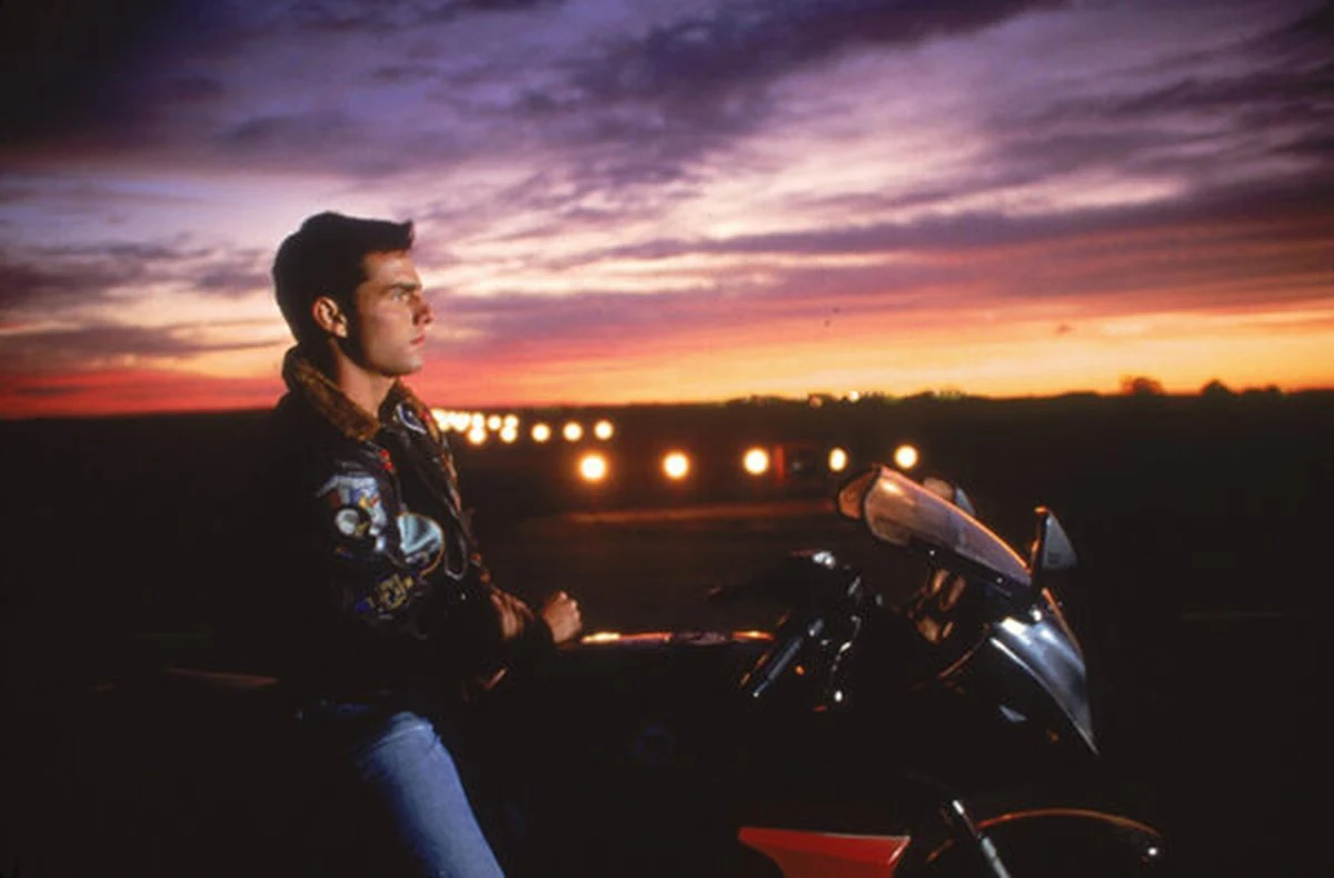 Maverick karakterini canlandıran Tom Cruise, pilot ceketini giymiş bir şekilde motosikletinin selesinde otururken, arka planda turuncu ve mor tonlardaki muhteşem gün batımı ile aydınlanan bir pistin uzaktan görünen ışıklarına doğru düşünceli bir ifadeyle bakıyor.