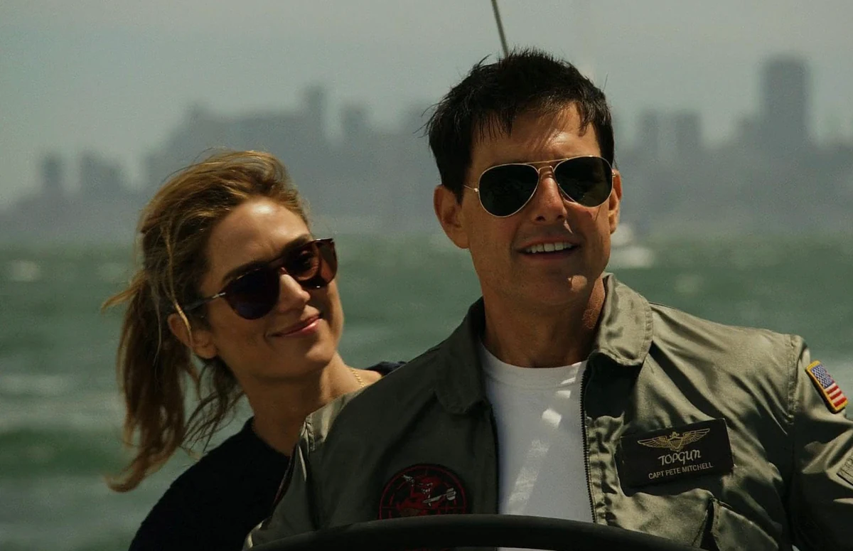 Pete Mitchell (Tom Cruise), Top Gun ceketi ve güneş gözlükleriyle bir yelkenlinin dümeninde hafifçe gülümserken, Penelope (Jennifer Connelly) de güneş gözlüklü ve at kuyruğu saçlarıyla arkasından ona sevgiyle bakıyor; bulanık şehir silüetiyle su üzerindeki huzurlu bir Top Gun Maverick sahnesi.