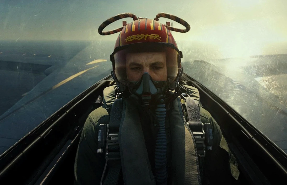 Top Gun: Maverick filminin ikonik kokpit sahnelerinden birinde, kırmızı sarı çizgili kaskında ROOSTER yazan ve oksijen maskesi takmış pilot, sisli gün batımı veya bulutlu bir gökyüzü altında uçağının kokpitinde ileriye odaklanmış şekilde görülüyor.