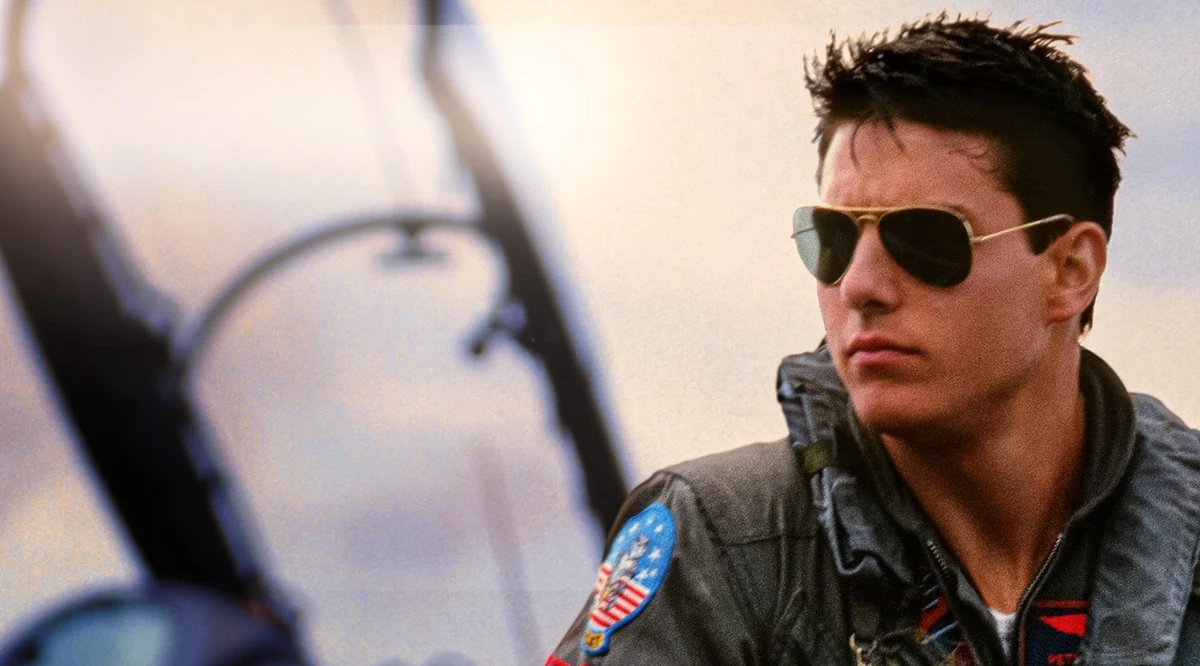 Genç Tom Cruise, Top Gun Maverick filmindeki ikonik rolüyle, omuzunda yamalı uçuş tulumu ve gözünde havacı gözlükleriyle, uçağının kokpitinde kararlı bir ifadeyle sağa doğru bakıyor.