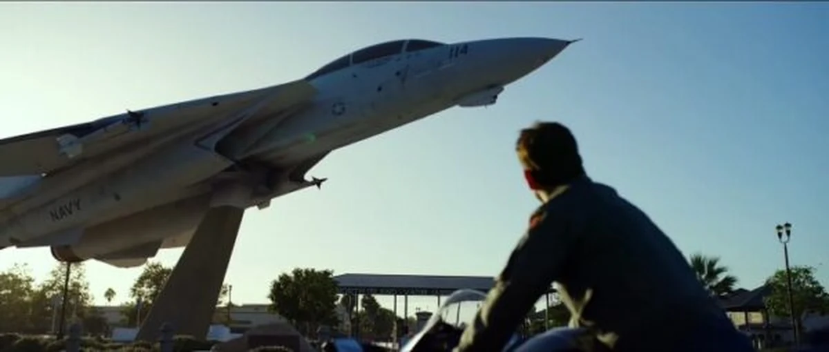 Maverick karakterini andıran bir motosikletli, güneşli bir açık hava sergisinde, üzerinde NAVY yazısı bulunan ve havada yükselir gibi bir kaide üzerinde duran gri bir F-14 Tomcat savaş uçağına dönüp bakıyor.