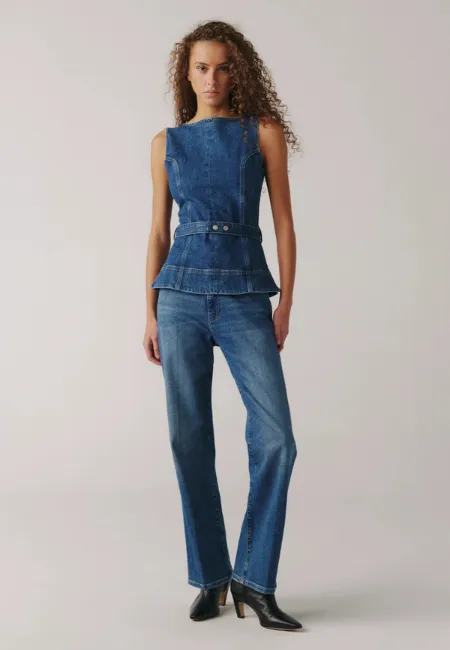 Mavi denim peplum kesim kolsuz üst ve düz paça jean pantolon giyen kadın model; siyah topuklu ayakkabıyla, sade açık renk stüdyo fonunda tam boy çekim.