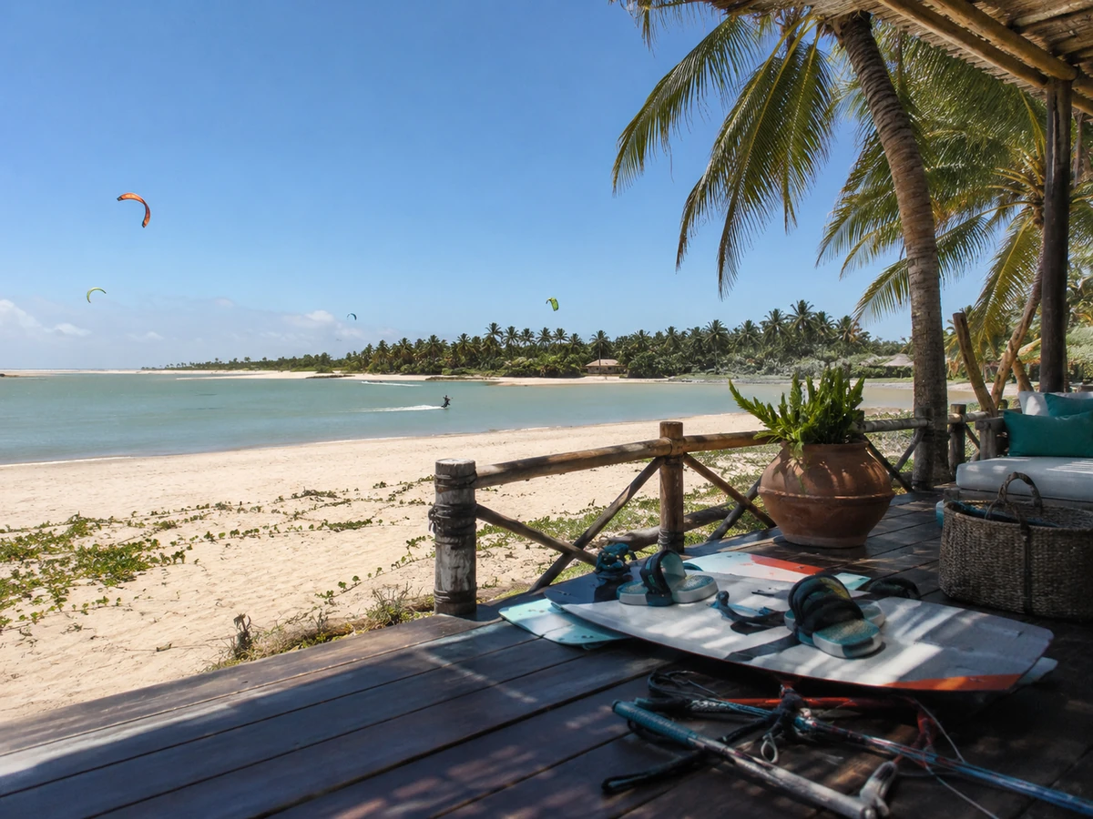 Kitesurf tahtası ve ekipmanlarının sergilendiği ahşap bir terastan görülen, Brezilya'nın Cumbuco sahillerine özgü palmiye ağaçlarıyla çevrili geniş kumlu plajda ve sığ turkuaz sularda birçok kişinin kitesurf yaptığı hareketli bir manzara.