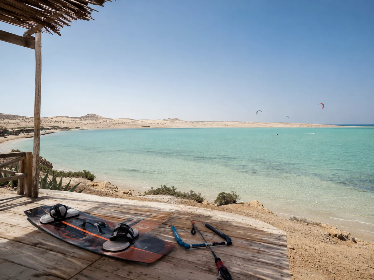 Ahşap bir platformda bekleyen bir kitesurf tahtası ve kontrol çubuğunun önünden görünen Dakhla'nın berrak turkuaz sularıyla çevrili kumsallı lagününde, uzaklarda çöl tepeleri yükselirken birkaç kitesurfçü kitesurf rotasında keyifli bir gün geçiriyor.