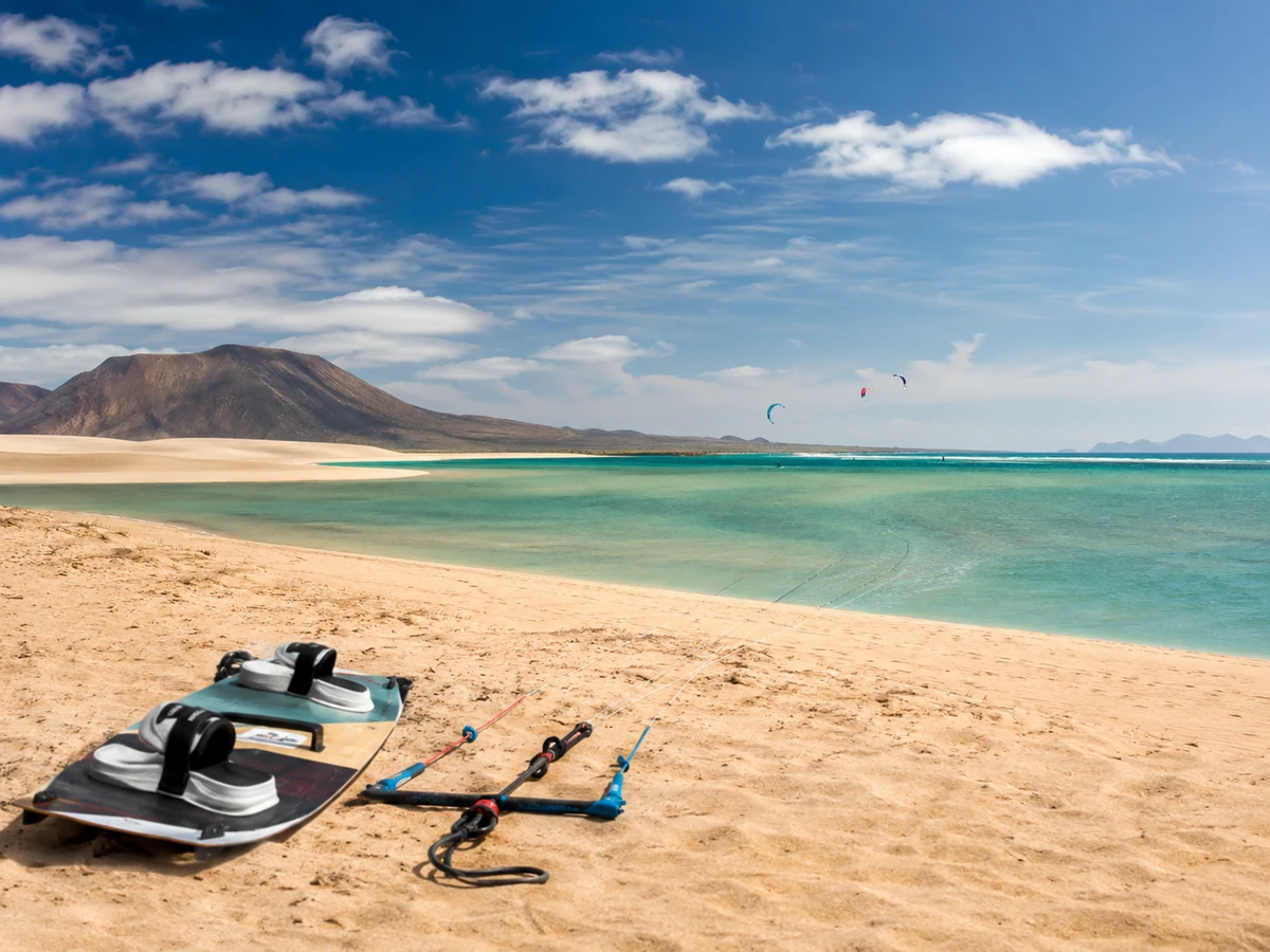 Fuerteventura'nın altın kumlu plajında, ön planda uzanmış bir kitesurf tahtası ve kontrol barı, turkuaz renkli denizde uzaklarda kitesurf yapan sporcular ve Kanarya Adaları'nın volkanik dağları ile bu popüler kitesurf rotasının doğal güzelliği vurgulanıyor.